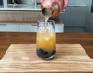 Passionfruit fruit tea being poured from a rose gold stainless steel shaker into a glass holding cooked dark purple Blueberry tapioca fruit pearls and ice