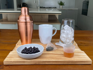 Rose gold cocktail shaker, cooked Blueberry tapioca fruit pearls, a tea bag, passionfruit fruit mix in a plastic scoop, glass with ice cubes and a white mug on a wooden dining table in a kitchen