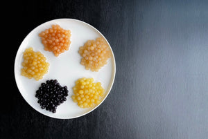 Cooked tapioca fruit pearls displayed on a white plate. Dark purple Blueberry, yellow Pineapple and Yuzu, off-white Lychee and pink Red Grapefruit pearls