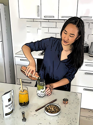 Marie in the kitchen pouring bright green Matcha tea from a rose gold shaker into a glass holding Blueberry tapioca fruit pearls, ice and milk