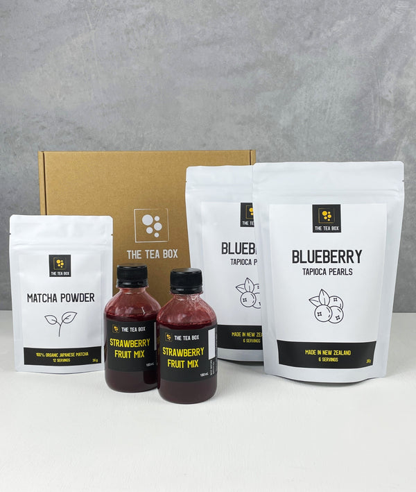 Contents of a 12 pack Strawberry Matcha Latte Bubble Tea Kit. Matcha powder and Blueberry pearls in pouches, 2 bottles of red Strawberry fruit mix, scoop, cardboard gift box