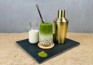 A glass of warm Matcha Latte Bubble Tea with Red Grapefruit tapioca fruit pearls and a silver stainless steel bubble tea straw. Displayed with gold cocktail shaker, vibrant green Matcha powder and a glass bottle of milk