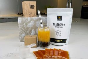 Passionfruit Bubble Tea Kit. Displayed with completed drink (bright orange tea and dark tapioca pearls), Passionfruit fruit mix sachets, pouch of blueberry pearls, gift wrapped box with Pohutukawa print and a Thank You card