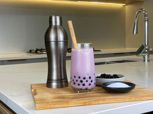 Rich purple Taro Bubble Tea with brown tapioca pearls and a reusable bamboo bubble tea straw. Displayed on a wooden chopping board with a dark grey stainless steel cocktail shaker, milk tea powder and cooked brown tapioca pearls in round ceramic dishes