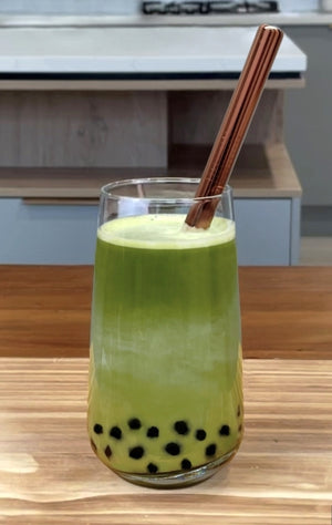 A glass of vibrant green warm Matcha Latte Bubble Tea with dark purple Blueberry tapioca fruit pearls and a rose gold stainless steel bubble tea straw