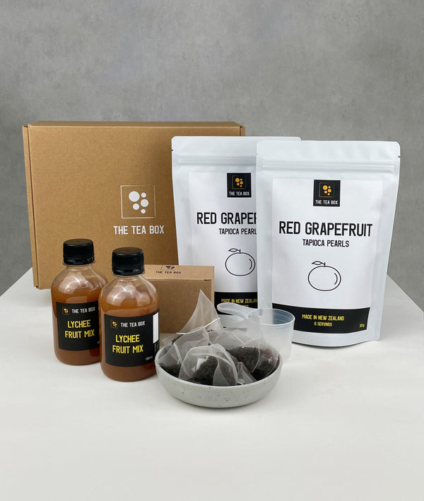 Contents of a 12 pack Lychee Bubble Tea Kit. Red Grapefruit pearls in pouches, bottles of Lychee fruit mix, tea bags, scoop and cardboard gift box