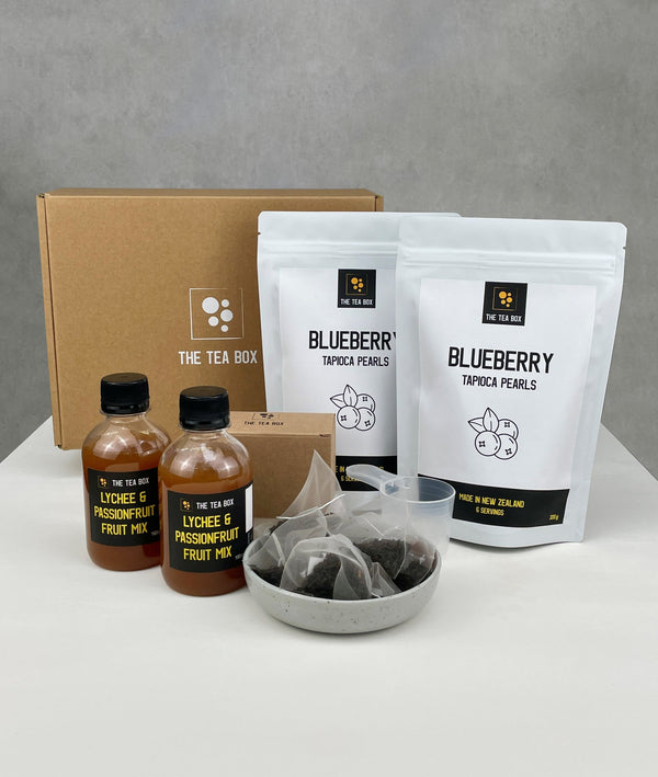 Contents of 12 pack Lychee and Passionfruit Bubble Tea Kit. Blueberry pearls in pouches, bottles of fruit mix, tea bags, scoop and cardboard gift box