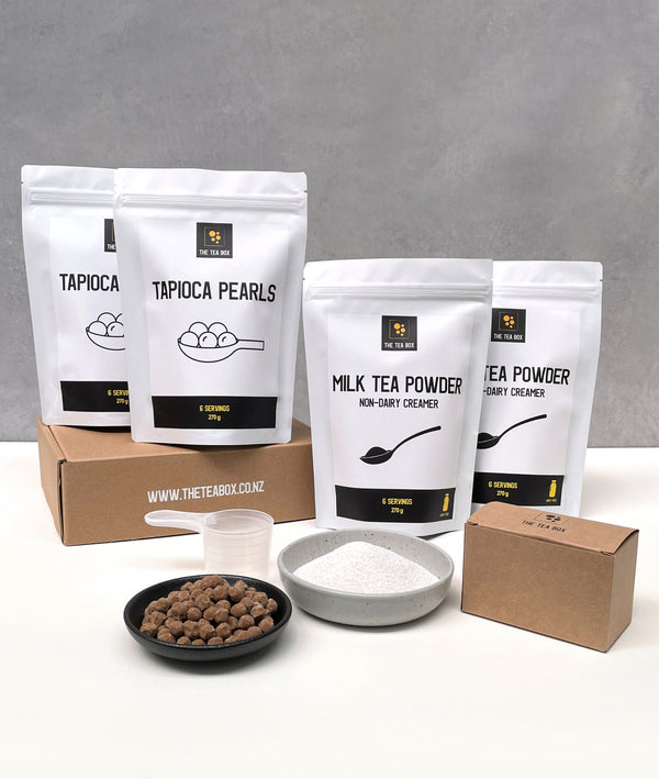 Contents of a 12 pack Pearl Milk Bubble Tea Kit. Milk tea powder and brown tapioca pearls in pouches, milk tea powder and uncooked brown tapioca pearls displayed in ceramic dishes, tea bags, scoop, cardboard gift box