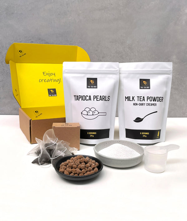 Contents of a 6 pack Pearl Milk Bubble Tea Kit. Milk tea powder and brown tapioca pearls in pouches, milk tea powder and uncooked brown tapioca pearls displayed in ceramic dishes, tea bags, scoop, cardboard gift box