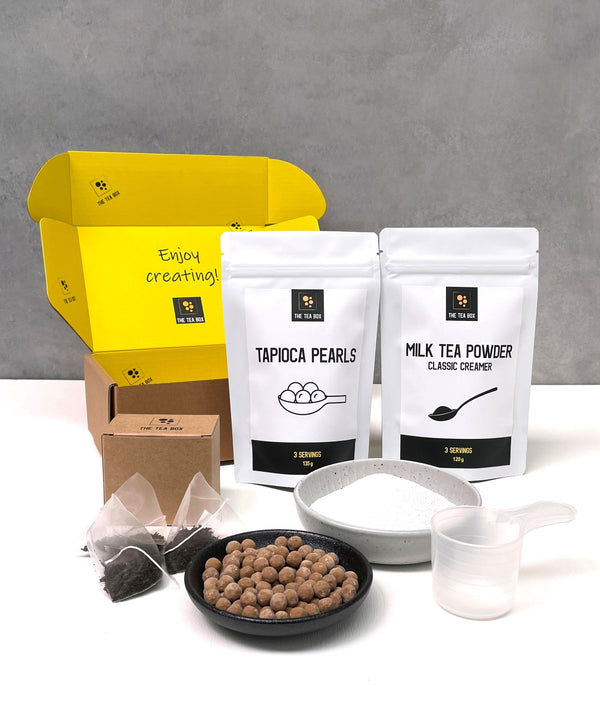 Contents of a 3 pack Pearl Milk Bubble Tea Kit. Milk tea powder and brown tapioca pearls in pouches, milk tea powder and uncooked brown tapioca pearls displayed in ceramic dishes, tea bags, scoop, cardboard gift box