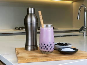 Rich purple Taro Bubble Tea with brown tapioca boba, bamboo straw. Displayed with dark grey cocktail shaker, taro powder