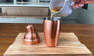 Brewed tea being poured from a white mug into a rose gold cocktail shaker