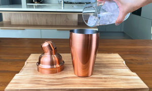 Half a glass of ice being added to a rose gold cocktail shaker