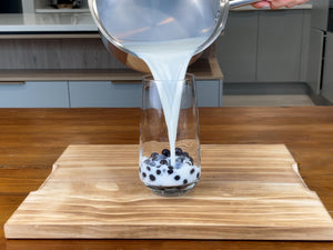 Two thirds of a glass of warmed milk being poured from a pot into a glass holding cooked Blueberry tapioca fruit pearls
