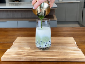 Vibrant green Matcha tea being poured from a gold stainless steel shaker into a glass holding milk and cooked, dark purple Blueberry tapioca fruit pearls