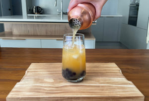 Passionfruit fruit tea being poured from a rose gold stainless steel shaker into a glass holding cooked dark purple Blueberry pearls and ice