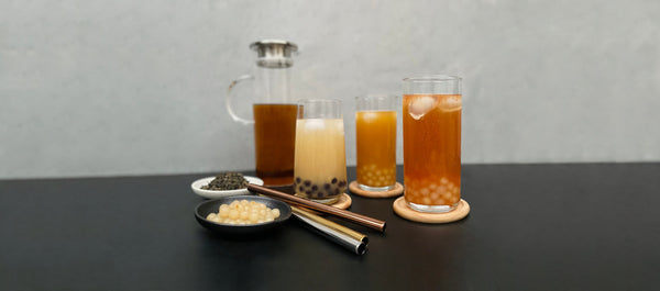3 brightly coloured fruit bubble teas with fruit pearls. Displayed with stainless steel straws, cooked yellow yuzu pearls, tea leaves and jug of brewed black tea