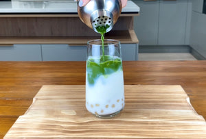 Vibrant green matcha tea being poured from a stainless steel shaker into a glass holding milk, cooked, pink-coloured Red Grapefruit pearls and ice