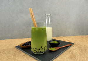 A glass of cold Matcha Latte Bubble tea with Blueberry pearls and reusable bamboo straw. Displayed with bright green matcha powder, tapioca pearls and milk