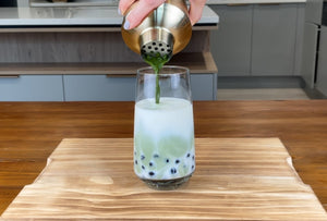 Vibrant green matcha tea being poured from a brushed gold stainless steel shaker into a glass holding milk and cooked dark purple Blueberry pearls