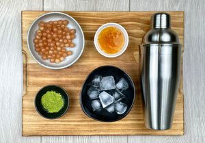 Stainless steel shaker with round ceramic dishes holding ice cubes, bright green Matcha powder, cooked pink, Red Grapefruit tapioca fruit pearls and yellow Yuzu fruit mix laying down on a wooden chopping board