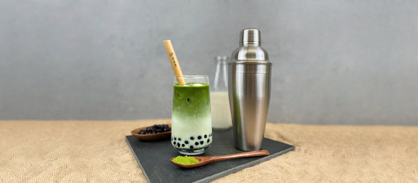 Bright green and white layered Matcha latte bubble tea with blueberry pearls, ice, bamboo bubble tea straw. Displayed with silver cocktail shaker, bright green matcha power, bottle of milk.