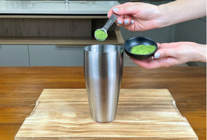 One teaspoon of bright green matcha powder being added to a stainless steel cocktail shaker