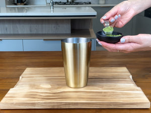 A black ceramic dish filled with bright green matcha powder. One teaspoon has been scooped out and is about to be added to a gold stainless steel cocktail shaker