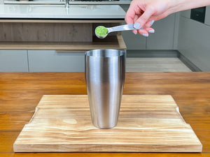 One teaspoon of bright green Matcha powder being added to a stainless steel cocktail shaker