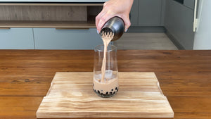 Light brown milk tea being poured from an antique grey stainless steel shaker into a glass holding cooked brown Tapioca pearls and ice