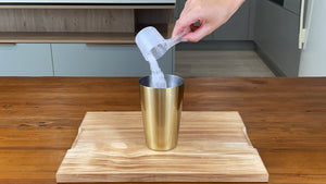 Milk tea powder being poured from a plastic scoop into a gold stainless steel cocktail shaker