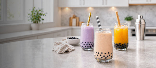 Three tall clear glasses of bubble tea with black tapioca pearls at the bottom, set on a light countertop in a bright kitchen. The drinks are Taro, Pearl Milk and Mango bubble teas each with a wide bubble straw, and a small bowl of black pearls sits to the left.