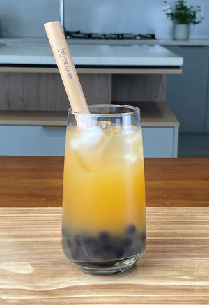 A glass of orange-coloured Passionfruit Bubble Tea with dark purple Blueberry tapioca fruit pearls, ice and a reusable bamboo bubble tea straw