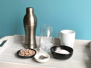 Antique grey stainless steel cocktail shaker displayed with round ceramic dishes holding milk tea powder, uncooked brown tapioca pearls and a tea bag. A glass with ice cubes and a white mug