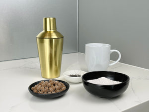 Gold stainless steel cocktail shaker with round ceramic dishes holding milk tea powder, uncooked brown tapioca pearls, a tea bag and a white mug