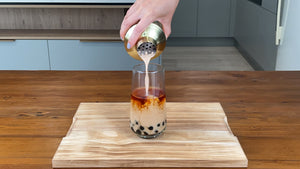 Milk tea being poured from a gold stainless steel shaker into a glass holding brewed tea and cooked brown tapioca pearls