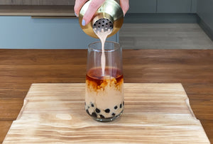 Light brown milk tea being poured from a gold stainless steel shaker into a glass holding tea and cooked brown tapioca pearls