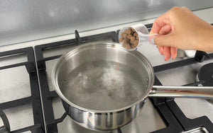 Uncooked brown tapioca pearls in a plastic scoop being added to a medium-sized pot of boiling water
