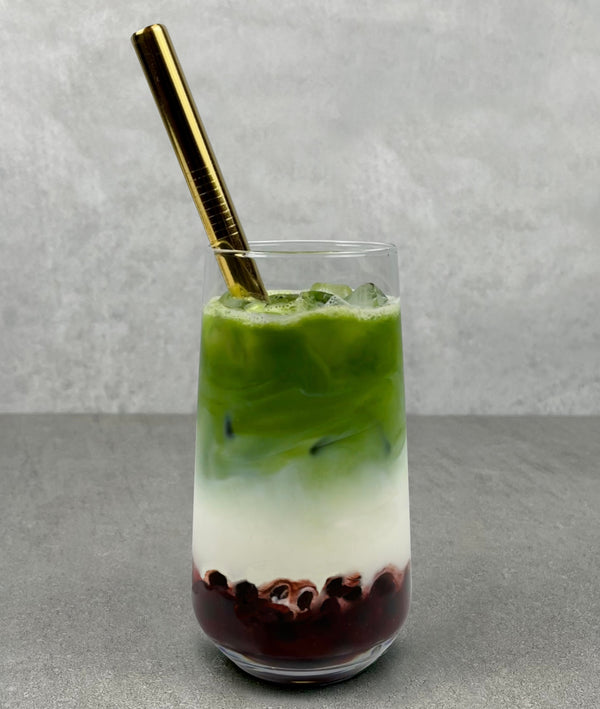 A glass of Strawberry Matcha Latte Bubble tea with Brown pearls, ice and a gold stainless steel bubble tea straw. Top layer is vibrant green matcha, bottom layer is dark red strawberry with pearls