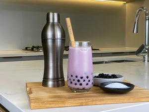 Rich purple Taro Bubble Tea with brown tapioca pearls and a reusable bamboo bubble tea straw. Displayed on a wooden chopping board with a dark grey stainless steel cocktail shaker, milk tea powder and cooked brown tapioca pearls in round ceramic dishes