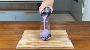 Bright purple taro milk tea being poured into a glass holding cooked brown tapioca pearls and ice cubes