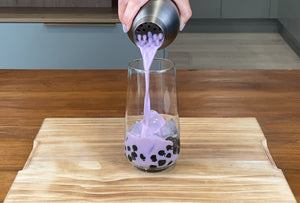 Purple Taro milk being poured from a dark grey antique shaker into a glass holding cooked brown tapioca pearls and ice cubes