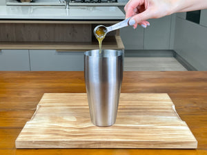 One teaspoon of yellow Yuzu fruit mix being added to a stainless steel cocktail shaker