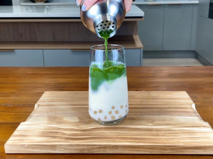 Vibrant green Matcha tea being poured from a stainless steel shaker into a glass holding milk, cooked pink-coloured Red Grapefruit tapioca fruit pearls and ice