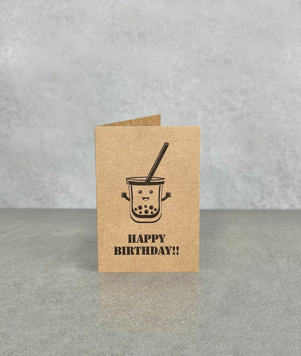 Happy Birthday card made of brown kraft card. Shows a cartoon bubble tea character smiling with arms waving. Card measures 70 x 100 mm when folded