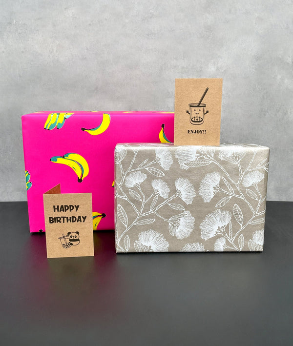 2 gift wrapped boxes. One with bright pink background covered in yellow and metallic green bananas, another with a silvery brown background covered in white pohutukawas. Along with two kraft card greeting cards