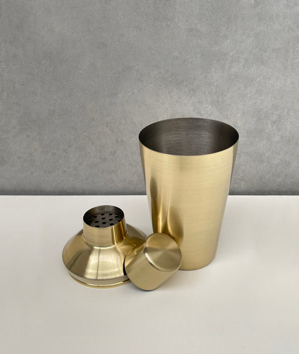 Unassembled brushed gold 3-piece shaker. Showing the cup, strainer and cap
