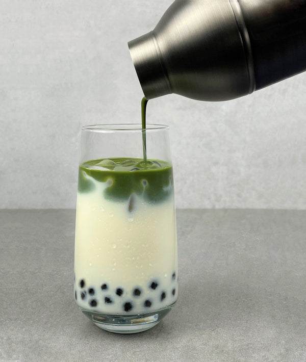 Green matcha tea being poured from a dark grey cocktail shaker into a glass. Creating a layer of vibrant green, atop ice, a layer of milk and Blueberry pearls