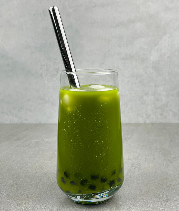 A glass of vibrant green Matcha Yuzu Bubble tea with Blueberry pearls, ice and a silver stainless steel bubble tea straw