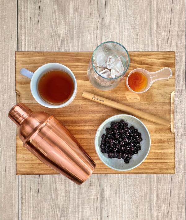 Rose gold shaker displayed with cooked Blueberry pearls, a bamboo straw, Lychee and Passionfruit fruit mix, a glass with ice and brewed tea in a cup